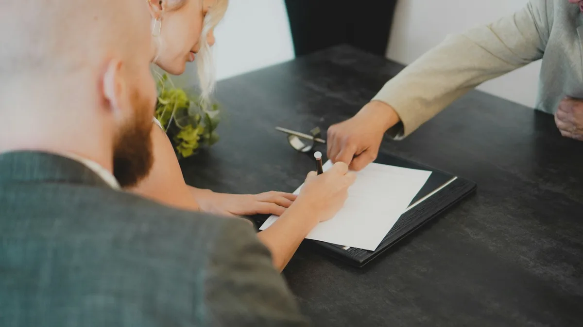 A couple seated at a desk signing a document with a pen, representing a U.S. buyer executing a deeded or right-to-use timeshare contract